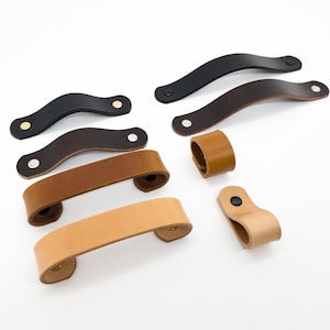 May include: A collection of leather cabinet pulls and handles in various colors, including black, brown, and tan. The hardware features a curved design and is shown against a white background. Some pulls have brass or silver rivets.