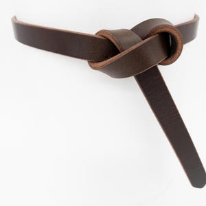 Può includere: Una cintura in pelle marrone scuro con un design annodato. La cintura è realizzata in un unico pezzo di pelle e presenta un nodo semplice ed elegante. La cintura è mostrata su uno sfondo bianco.