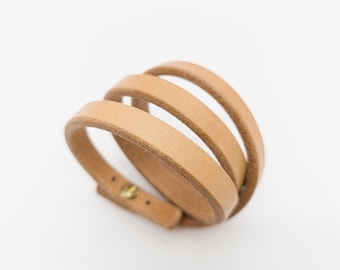 May include: A light brown leather wrap bracelet with three bands. The bracelet has a simple design with a brass-colored clasp. The bands are approximately 0.5 inches wide.