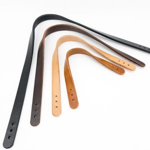 Könnte beinhalten: Eine Auswahl an Leder-Taschengriffen in verschiedenen Farben: Schwarz, Dunkelbraun, Mittelbraun, Beige und Hellbraun. Jeder gebogene Griff hat drei vorgebohrte Löcher zur Befestigung. Auf weißem Hintergrund angeordnet.