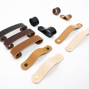 Peut inclure: Une collection de poignées et de tirettes de meubles en cuir de différentes couleurs, dont noir, marron et beige. La quincaillerie présente des designs incurvés et en boucle, avec des rivets métalliques pour la fixation.