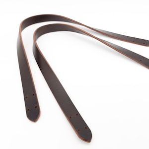 Puede incluir: Dos correas de bolso de cuero marrón oscuro con extremos redondeados y agujeros pretaladrados para la fijación. Las correas están curvadas y se apoyan sobre un fondo blanco, mostrando su textura suave y su rico color.