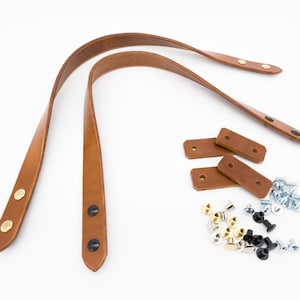 May include: A set of brown leather bag handles with gold and black rivets, along with matching leather reinforcement pieces and various screws and rivets, all on a white background. Ideal for DIY projects.