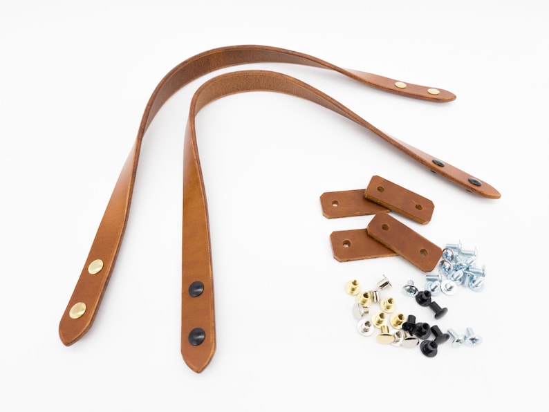 May include: A set of brown leather bag handles with gold and black rivets, along with matching leather reinforcement pieces and various screws and rivets, all on a white background. Ideal for DIY projects.