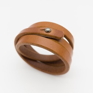 Könnte beinhalten: Ein braunes Lederarmband mit Metallknopfverschluss. Das Armband besteht aus mehreren Lederschichten und ist zum Wickeln um das Handgelenk konzipiert. Das Leder hat ein natürliches, strukturiertes Aussehen.