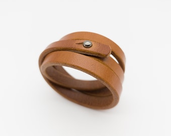 May include: A brown leather wrap bracelet with a metal stud closure. The bracelet is made of multiple layers of leather and is designed to wrap around the wrist. The leather has a natural, textured appearance.