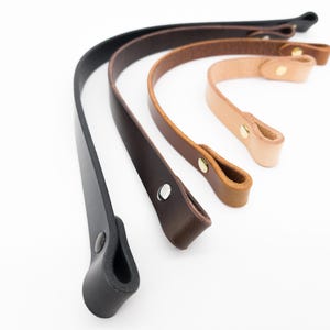 Può includere: Quattro manici per borse in pelle, in varie tonalità di marrone e nero, sono disposti diagonalmente su uno sfondo bianco. Ogni manico ha un'asola a un'estremità ed è fissato con un rivetto metallico. I manici variano in lunghezza.