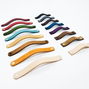 May include: A selection of leather drawer pulls in a range of colours, such as red, green, and brown. Each pull features a curved design and is fastened with metal rivets. The pulls are displayed against a white backdrop.