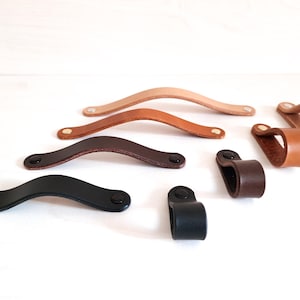 以下が含まれることがあります： さまざまな色の革製引き出しプルとキャビネットハンドルのコレクション。ベージュ、茶色、黒の色合いの湾曲したプルと、ループスタイルのハンドルが含まれています。各ピースは黒いハードウェアで固定されています。