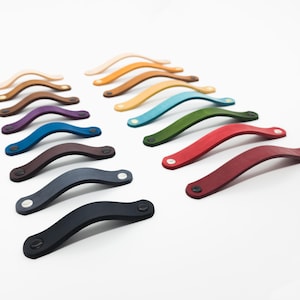 Peut inclure: Une collection de poignées de meubles en cuir coloré, dans des teintes variées, dont le marron, le bleu, le vert et le rouge. Chaque poignée est incurvée et fixée avec deux petits rivets argentés, offrant une esthétique moderne.