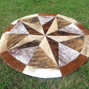 Pode incluir: Tapete redondo de pele de vaca com um desenho de estrela. O tapete apresenta um padrão de estrela central em tons de castanho, bege e branco, rodeado por uma borda de manchas castanhas e brancas. O tapete está sobre a relva verde.