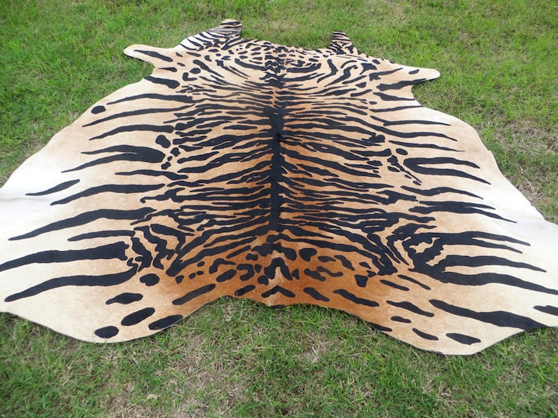 May include: A large, tan and black cowhide rug with a tiger stripe pattern. The rug is laid out on green grass, showcasing the natural hide's shape and design. The black stripes are bold against the tan background.