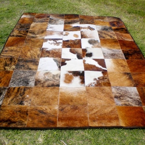 Puede incluir: Una alfombra de piel de vaca patchwork con un patrón de tablero de ajedrez en tonos marrones, blancos y negros. La alfombra está hecha de piezas individuales de piel de vaca cosidas juntas para crear un aspecto único y rústico.