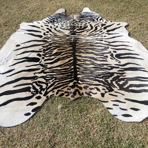 May include: A cowhide rug with a striking tiger stripe pattern in black and cream. The rug is irregularly shaped, with a natural edge, and lies on a grassy surface. The design is bold and eye-catching, suitable for a living room or bedroom.