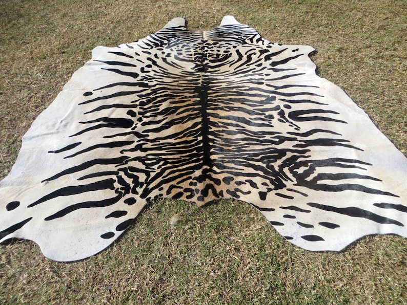 May include: A cowhide rug with a striking tiger stripe pattern in black and cream. The rug is irregularly shaped, with a natural edge, and lies on a grassy surface. The design is bold and eye-catching, suitable for a living room or bedroom.