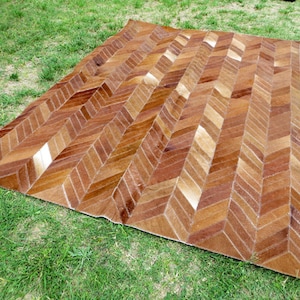May include: A rectangular rug with a herringbone pattern in shades of brown and tan. The rug is made of a natural material and is placed on a bed of green grass. The rug's design creates a visual texture.