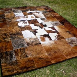 May include: A patchwork rug made of cowhide with brown, white and black patches. The rug is laid out on a green grassy surface.