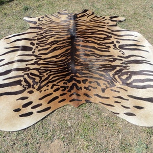 May include: A cowhide rug with a tiger stripe pattern. The rug features a light tan background with dark brown stripes and spots, creating a striking animal print design. The rug is irregularly shaped, with a natural edge.