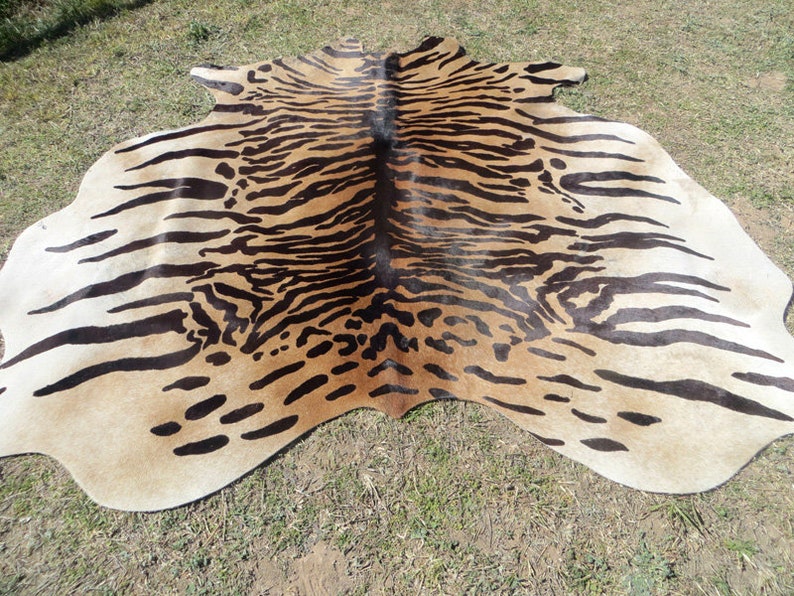 May include: A cowhide rug with a tiger stripe pattern. The rug features a light tan background with dark brown stripes and spots, creating a striking animal print design. The rug is irregularly shaped, with a natural edge.
