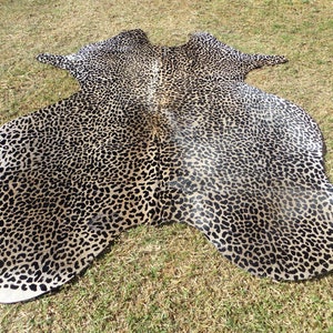 May include: A large, brown and black cowhide rug with a leopard print pattern. The rug is laid out on a grassy surface.