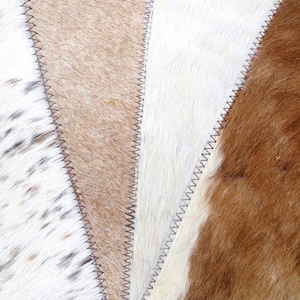 May include: A close-up of a patchwork rug made of different colored cowhide. The rug is made of four different pieces of cowhide, each with a different pattern. The rug is sewn together with a zig-zag stitch.