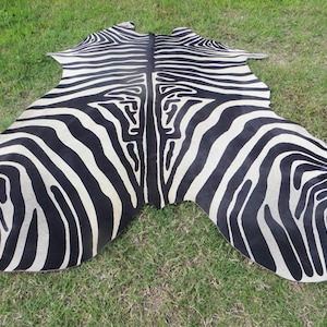 May include: Black and white zebra hide rug with a natural, slightly textured surface. The rug is laid out on a green grassy surface.
