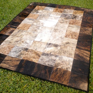 May include: A patchwork cowhide rug with a black border. The rug is made of different shades of brown, white, and gray cowhide.