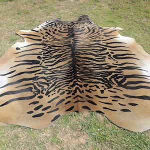 Puede incluir: Una alfombra de piel de vaca marr&oacute;n y negra con un estampado de tigre. La alfombra est&aacute; colocada sobre una superficie cubierta de hierba.