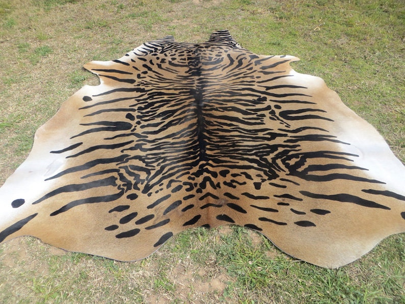 Puede incluir: Una alfombra de piel de vaca marr&oacute;n y negra con un estampado de tigre. La alfombra est&aacute; colocada sobre una superficie cubierta de hierba.