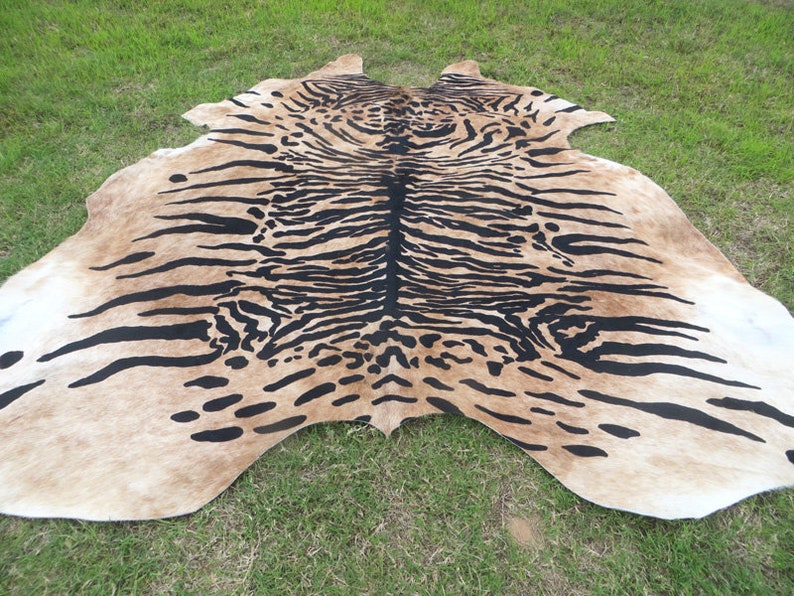 May include: A cowhide rug with a tiger stripe pattern. The rug features a tan background with black stripes, creating a striking contrast. The rug is laid out on green grass, showcasing its natural shape and texture.