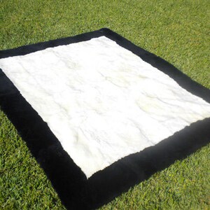Puede incluir: Una alfombra de piel sintética blanca con un borde negro. La alfombra está colocada sobre una superficie de hierba verde.