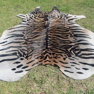 May include: A cowhide rug with a tiger stripe pattern. The rug features a mix of tan, black, and white colors. The rug is lying on a green grassy surface.
