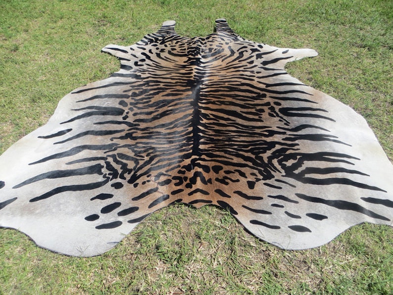 May include: A cowhide rug with a tiger stripe pattern. The rug features a mix of tan, black, and white colors. The rug is lying on a green grassy surface.