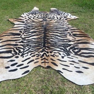 May include: A cowhide rug with a tiger stripe pattern. The rug features a beige background with black stripes and spots, creating a striking animal print design. The rug is irregularly shaped, with rounded edges and a natural appearance.