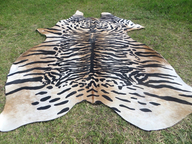 May include: A cowhide rug with a tiger stripe pattern. The rug features a beige background with black stripes and spots, creating a striking animal print design. The rug is irregularly shaped, with rounded edges and a natural appearance.