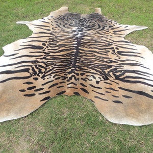May include: A cowhide rug with a tiger stripe pattern. The rug features a tan background with black stripes and spots, and white edges. The rug is laid out on green grass.