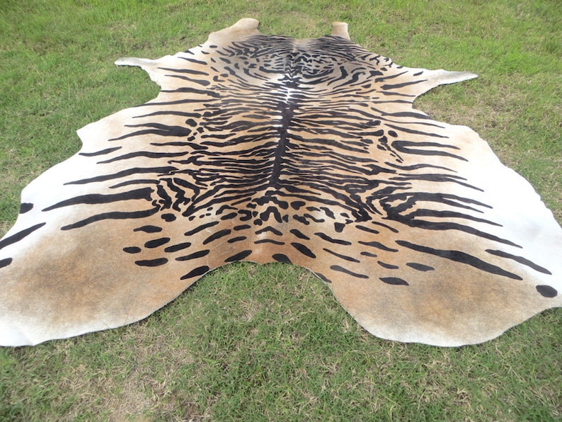 May include: A cowhide rug with a tiger stripe pattern. The rug features a tan background with black stripes and spots, and white edges. The rug is laid out on green grass.