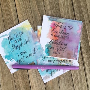 May include: Four watercolour cards with inspirational quotes from the bible. The cards feature a watercolour background in shades of blue, green, pink, and yellow. The quotes are written in black calligraphy. The quotes are: "The Lord is my Shepherd, I shall not want", "He makes me lie down in green pastures, He leads me beside still waters", "They comfort me", and "Forever".