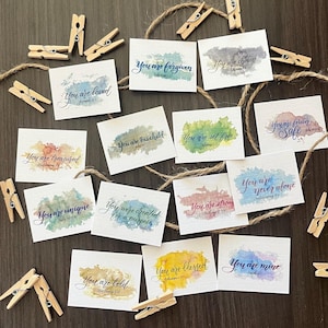 May include: A collection of small, square cards with watercolor backgrounds and inspirational phrases in elegant script. The cards are arranged with twine and wooden clothespins, featuring messages like "You are loved" and "You are forgiven."