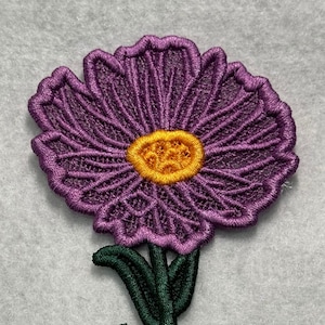 May include: Purple and green embroidered flower applique with a yellow center. The flower has a delicate, lacy design.