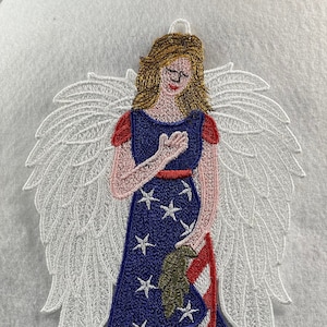 May include: Embroidered angel ornament with white wings, a blue dress with white stars and red and white stripes, and a gold trim. The angel is holding a green olive branch in her hand.