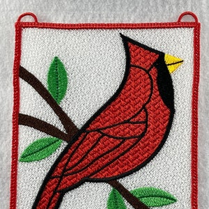 May include: Embroidered patch featuring a red cardinal perched on a branch with green leaves. The cardinal has a black beak and a yellow spot on its head. The patch is surrounded by a red border.