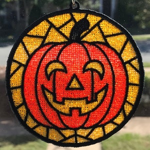 May include: A round, orange and yellow pumpkin-shaped window hanging with a black spiderweb design. The pumpkin has a carved jack-o'-lantern face.