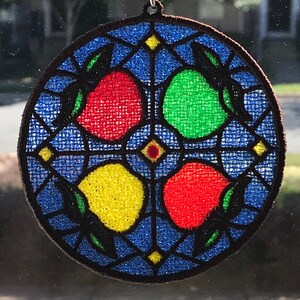 May include: A round, blue, embroidered, stained glass-style ornament with four fruit designs. The fruit are red, green, yellow, and red. The ornament is hanging from a suction cup.