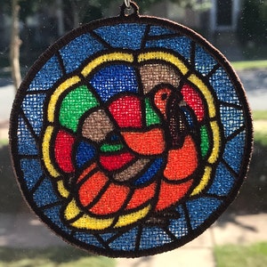May include: A round, stained glass-style ornament featuring a colorful turkey design. The turkey is outlined in black and has a red head, orange body, and green and blue accents. The ornament is surrounded by a brown border.