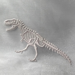May include: White embroidered dinosaur skeleton patch. The dinosaur is standing on all fours with its tail curled up. The patch is perfect for adding a touch of whimsy to any project.