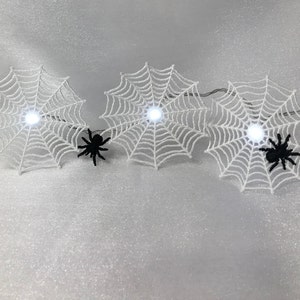 May include: Three white spiderwebs with black spiders and white lights strung together on a white background.