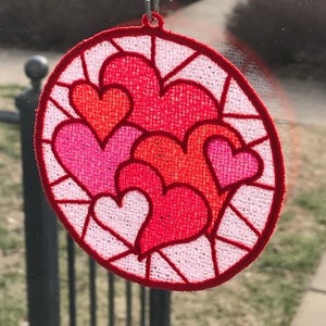 May include: A round, red, embroidered heart-shaped ornament with a white background and a white hanging loop. The ornament features multiple layers of hearts in shades of pink and red.