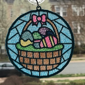 May include: A blue and white stained glass Easter basket ornament with a pink bow and colorful eggs inside. The basket is made of a woven material and has a handle. The ornament is hanging from a window.