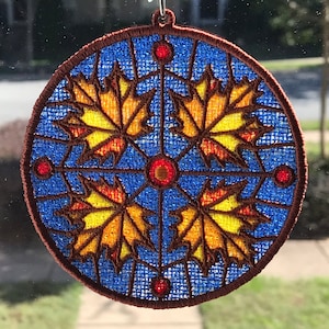 May include: A round, blue, brown, and yellow embroidered ornament with a design of four stylized maple leaves. The ornament is hanging from a window.